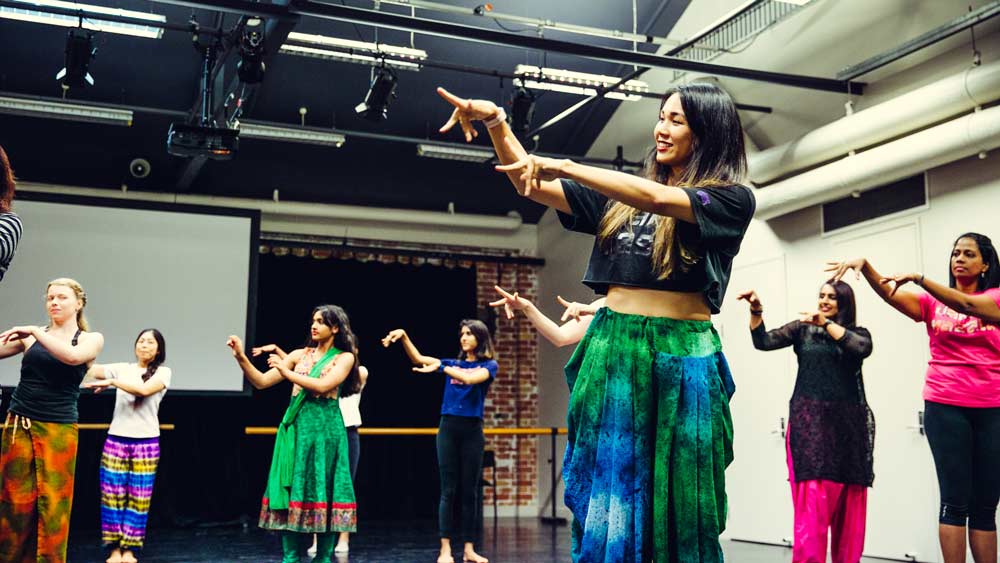An image of a pretty woman teaching a group of women dancers with their arms lifted up and their fingers outstretched classes